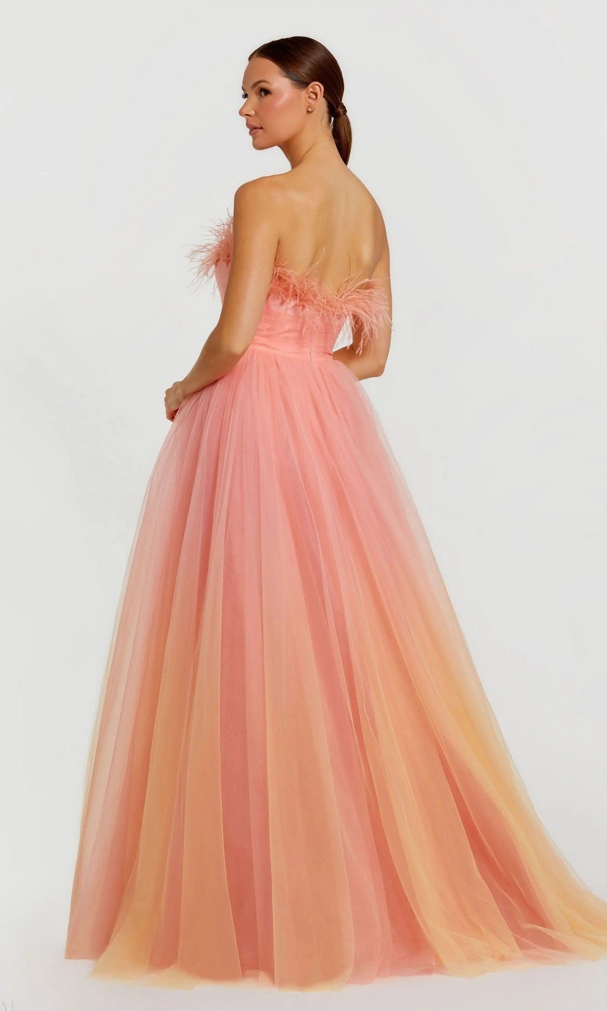 A woman poses in the Long Formal Dress 20557 by Mac Duggal, a strapless peach-pink tulle ball gown with ombre feather detailing on the bodice, standing slightly turned against a plain background.